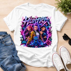 May include: A white short-sleeved t-shirt featuring a vibrant graphic print of three stylized characters and a blue cat. The design includes the text "HUNTRIX" in a bold, angular font, surrounded by neon pink and blue lightning, stars, and graffiti-style elements.