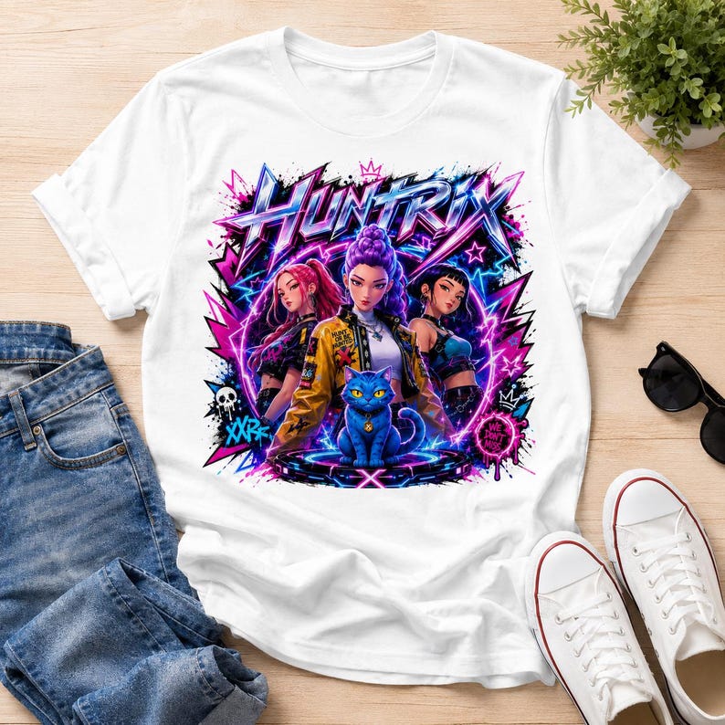 May include: A white short-sleeved t-shirt featuring a vibrant graphic print of three stylized characters and a blue cat. The design includes the text "HUNTRIX" in a bold, angular font, surrounded by neon pink and blue lightning, stars, and graffiti-style elements.