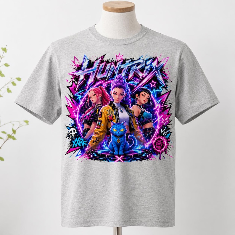 May include: A heather gray short-sleeve t-shirt featuring a vibrant graphic print with the text "HUNTRIX". The design displays three anime-style characters and a central blue cat sitting on a glowing platform, surrounded by neon pink and blue lightning.