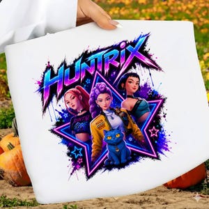 May include: A white t-shirt featuring a graphic print of three characters and a blue cat inside a neon star. The text "HUNTRIX" appears in a stylized, vibrant blue and pink font above the characters. The design includes purple and blue paint splatters.