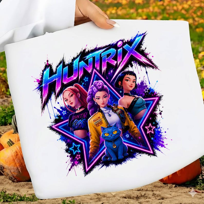 May include: A white t-shirt featuring a graphic print of three characters and a blue cat inside a neon star. The text "HUNTRIX" appears in a stylized, vibrant blue and pink font above the characters. The design includes purple and blue paint splatters.