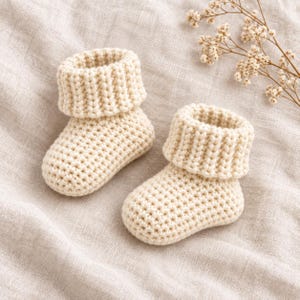 May include: A pair of cream-coloured baby booties crafted with a chunky knit texture. Each bootie features a thick, folded cuff at the top and a rounded toe design. The footwear is made from a soft yarn, showcasing a consistent stitch pattern throughout.