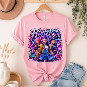 May include: A light pink t-shirt featuring a graphic print with the text HUNTRIX. The design displays three stylized characters and a blue cat surrounded by vibrant purple and pink lightning bolts, stars, and graffiti-style elements.