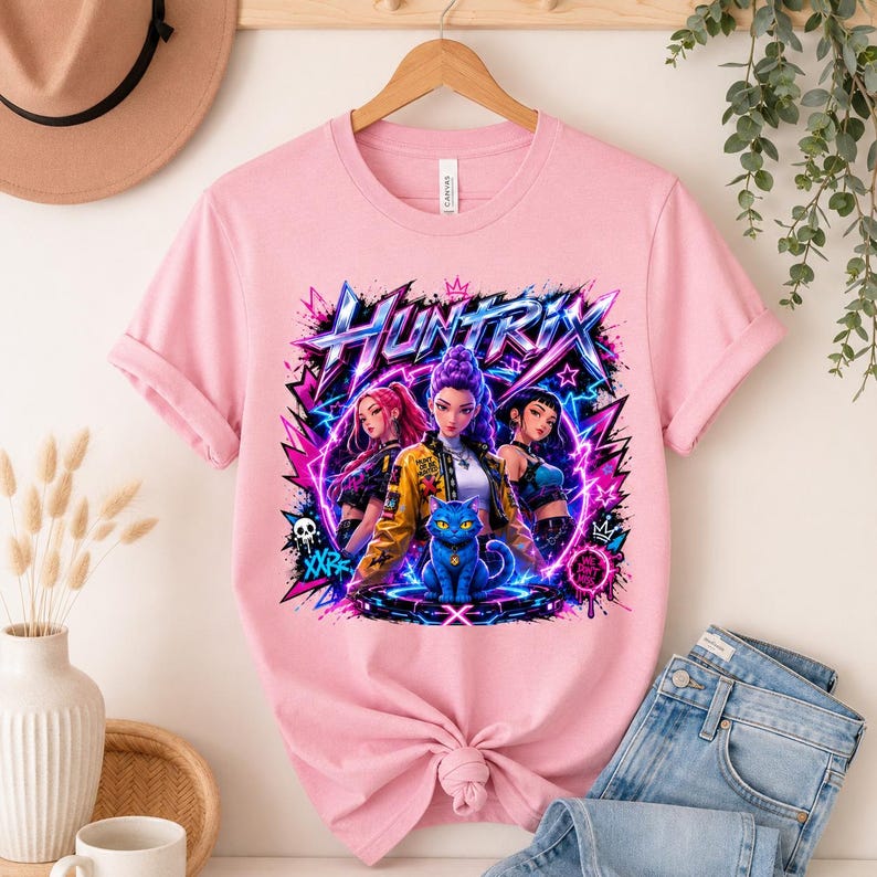 May include: A light pink t-shirt featuring a graphic print with the text HUNTRIX. The design displays three stylized characters and a blue cat surrounded by vibrant purple and pink lightning bolts, stars, and graffiti-style elements.