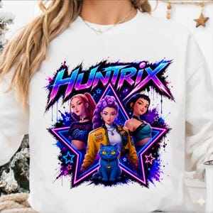 May include: A white sweatshirt featuring a graphic print with the text HUNTRIX in a stylized neon font. The design includes three illustrated characters and a blue cat centered within a glowing star shape, surrounded by vibrant purple and blue splashes.