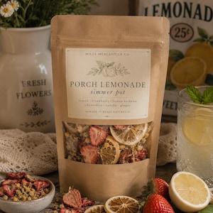 Könnte beinhalten: Ein brauner Papierbeutel mit dem Etikett Mills Mercantile Co. Porch Lemonade simmer pot, gefüllt mit getrockneten Zitronenscheiben, Erdbeeren und Kamillenblüten. Das Etikett listet Zutaten wie Zitrone, Erdbeere, Zitronenverbene, Kamille, Vanille und Ingwer auf.
