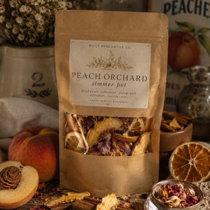 Puede incluir: Una bolsa de papel marrón etiquetada como Mills Mercantile Co. Peach Orchard simmer pot que contiene melocotón seco, caléndula, piel de naranja, canela, vainilla y rosa. La ventana transparente muestra los ingredientes secos del interior.