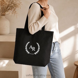 Peut inclure: Un sac fourre-tout en toile noire orné d'un motif de couronne de laurier circulaire blanc avec les lettres AR centrées à l'intérieur. Le sac possède deux bandoulières et est fabriqué dans un tissu noir uni au design épuré et simple.