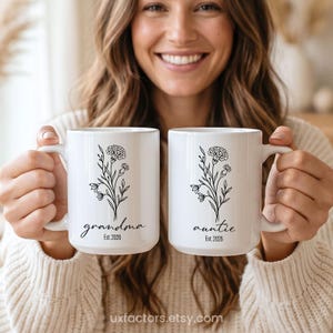 May include: Two white ceramic mugs featuring a black line art illustration of a carnation flower. The left mug has the text "grandma" and "Est. 2026" printed below the flower, while the right mug displays "auntie" and "Est. 2026" in black script.