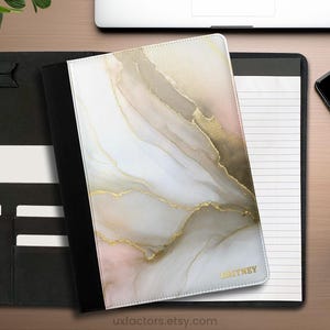 May include: A black portfolio folder featuring a white, beige, and soft pink marble-patterned cover with gold-coloured veining accents. The cover includes the name BRITNEY printed in gold lettering. The interior contains a lined notepad and slots.