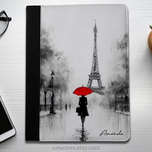 May include: A rectangular tablet or notebook cover featuring a black and white watercolour-style illustration of the Eiffel Tower in Paris. A figure walks towards the tower holding a bright red umbrella. The name Amanda is printed in black script.