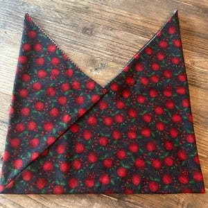 May include: A triangular fabric bandana featuring a repeating pattern of small red apples with green leaves and stems set against a dark black background. The fabric is folded into a sharp point at the top, displaying the printed textile design clearly.