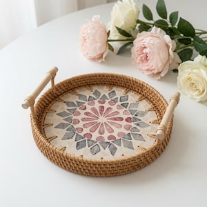 Puede incluir: Una bandeja redonda de ratán tejido con dos asas de madera clara en los lados opuestos. La base de la bandeja muestra un patrón decorativo incrustado con un diseño floral rosa central rodeado de formas geométricas grises y blancas.