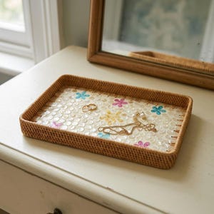 May include: A rectangular woven rattan tray featuring a base inlaid with small, circular white shell pieces and scattered floral motifs in blue, pink, and yellow. Two gold-toned rings and a delicate gold chain necklace rest on the tray surface.