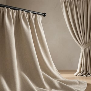 Peut inclure: Une paire de rideaux beige pleine longueur en tissu texturé. Un rideau pend librement depuis une tringle en métal noir, tandis que l'autre est rassemblé et maintenu par une embrasse assortie, créant des plis verticaux et un drapé.