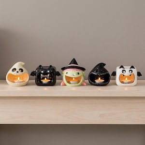 Puede incluir: Cinco portavelas de cerámica con forma de personajes de Halloween, incluyendo un fantasma crema, un murciélago negro, una bruja verde con sombrero rosa, un fantasma negro y un murciélago blanco, cada uno con una vela encendida en la boca.