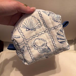 May include: A white quilted pouch featuring a blue line-art pattern of various seashells arranged in a grid. The pouch has a white zip closure along the top and small blue fabric tabs on the sides. The fabric has a textured, stitched appearance.