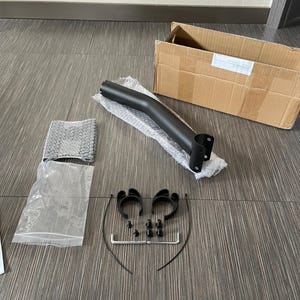 Könnte beinhalten: Ein mattschwarzer Metallstützarm, zwei kreisförmige schwarze Kunststoffklemmen, vier kleine schwarze Schrauben, zwei L-förmige Inbusschlüssel und zwei schwarze Kabelbinder. Die Komponenten liegen neben einem Versandkarton und Luftpolsterfolie.