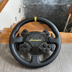 May include: A black racing steering wheel featuring a yellow center marker at the top, a carbon fiber center plate with the Lamborghini logo in yellow, and multiple integrated control buttons, dials, and paddle shifters on the rear side.