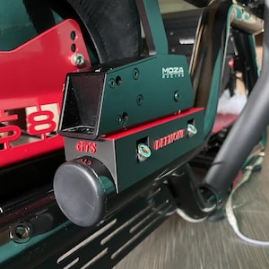 Peut inclure: Un frein à main de simulation de course Moza Racing vert foncé et noir avec des accents rouges. L'appareil porte l'inscription 'MOZA RACING' sur le côté, 'GTS' sur l'avant et 'DEEMONE' en lettres rouges sur le côté, monté sur un cadre en métal noir.