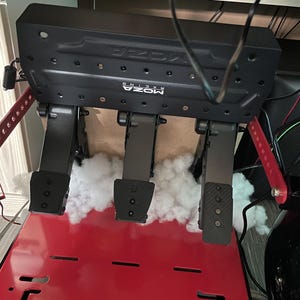 May include: A black Moza Racing pedal set featuring three pedals with textured surfaces, mounted on a black base with a red metal floor plate. White synthetic fiber padding is tucked behind the pedals, and black cables are visible above.