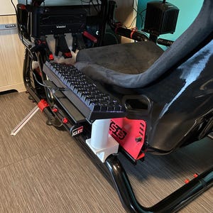Puede incluir: Un cockpit de simulador de carreras negro que incluye un asiento tipo cubo con tapicería gris oscuro, un teclado mecánico negro montado en un soporte blanco, un marco de metal rojo, una palanca de cambios y pedales marca Moza en negro y rojo.