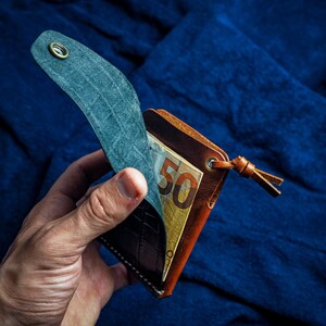 Op de afbeelding: Een bruine lederen portemonnee met een getextureerde donkerblauwe flap voorzien van een metalen drukknoopsluiting. De portemonnee bevat een biljet van vijftig euro en heeft een klein leren treklipje aan een metalen oogje aan de zijkant.
