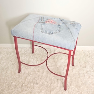 May include: A rectangular stool featuring a seat upholstered in light blue denim fabric with visible seams, a back pocket, and a small red and white checkered cloth tucked inside. The base is constructed from four curved red metal legs.