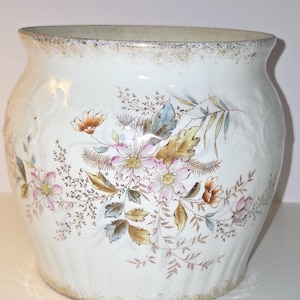 May include: A white ceramic planter with a wide, rounded body and a flared rim. The exterior features a delicate floral pattern in shades of pink, brown, and blue, with embossed scrollwork details and a weathered gold-toned trim on the edges.