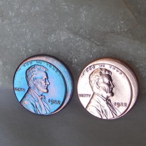 Może przedstawiać: Dwie okrągłe metalowe monety z profilem Abrahama Lincolna, napisem IN GOD WE TRUST, LIBERTY oraz rokiem 1922. Jedna moneta ma metaliczne niebiesko-fioletowe opalizujące wykończenie, a druga standardowe wykończenie w kolorze miedzi.