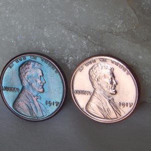 May include: Two 1917 Lincoln wheat pennies displayed side by side. The left coin features a distinct iridescent blue and purple patina, while the right coin exhibits a bright, polished copper finish. Both coins show the profile of Abraham Lincoln.