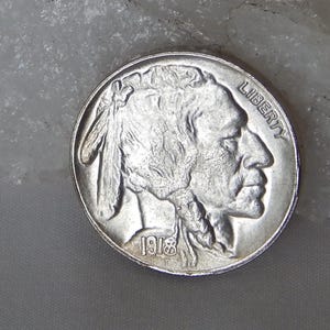 May include: A silver-colored 1918 Buffalo nickel coin featuring the profile of a Native American figure facing right. The word LIBERTY is inscribed along the upper right edge, and the year 1918 is clearly visible at the bottom of the coin face.