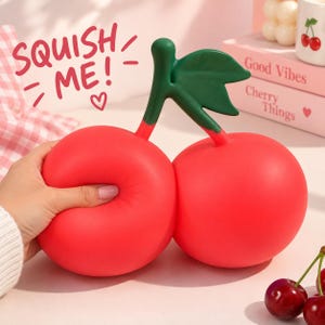 Może przedstawiać: Czerwona zabawka typu squishy w kształcie wiśni z zieloną łodyżką i listkiem. Zabawka posiada napis SQUISH ME! różowymi literami. Powierzchnia jest gładka i matowa, widać dłoń naciskającą jedną z dwóch okrągłych części wiśni.