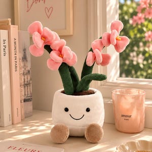 Peut inclure: Une peluche représentant un pot blanc avec un visage souriant et deux petits pieds marron, contenant deux tiges en velours côtelé vert surmontées de fleurs en tissu rose tendre. Le pot a des yeux brodés noirs et une bouche incurvée.