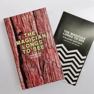 Puede incluir: Un juego de cartas de tarot de Twin Peaks con dos elementos rectangulares. Uno tiene un patrón de veta de madera roja con el texto THE MAGICIAN LONGS TO SEE. El segundo es negro con patrones de chevrón blancos y el texto THE MAGICIAN LONGS TO SEE.
