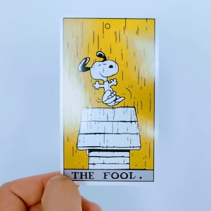 Puede incluir: Una carta de tarot rectangular con fondo amarillo con líneas verticales y un personaje de perro blanco saltando sobre una caseta de perro blanca. El texto THE FOOL está impreso en letras mayúsculas negras en la parte inferior dentro de un borde blanco.