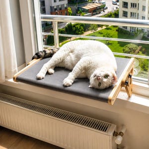 May include: A white cat with a grey-tipped tail lying on a rectangular grey fabric cat perch. The perch features light-coloured wooden side supports and is mounted to a window frame with suction cups, positioned above a white metal radiator.