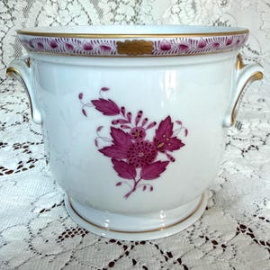 May include: A white porcelain cachepot featuring a magenta floral motif on the front. The rim and handles are accented with gold trim, and a decorative magenta leaf pattern encircles the upper edge of the vessel, which has two side handles.