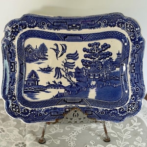 May include: A rectangular ceramic platter featuring a blue and white willow pattern. The design depicts a landscape with a bridge, a pagoda, trees, and two birds in flight. The border is decorated with intricate geometric and floral motifs.