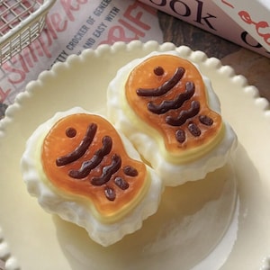 May include: Two fish-shaped cakes with a golden-brown exterior and dark brown detailing on a scalloped-edge cream-colored plate. The cakes feature a smooth, glossy finish with a distinct fish silhouette, including a circular eye and wavy fin lines.