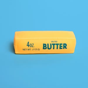 May include: A rectangular block of yellow salted butter with dark blue text printed on the front. The text reads 4oz. NET WT. (113 G) SALTED BUTTER. The block has smooth, rounded edges and a solid, uniform yellow colour throughout the surface.
