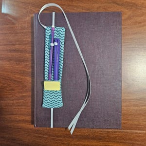 May include: A dark purple hardcover book featuring a teal and black chevron patterned elastic pen holder attached to the cover. The holder contains a purple pen, a teal pen, and a small yellow sticky note pad, secured by a white elastic band.