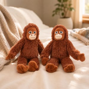 Op de afbeelding: Twee pluche orang-oetan knuffels met ruige, donkerbruine vacht, lichtbruine gezichten en geborduurde gelaatstrekken. Elk knuffeldier heeft lange armen, ronde oren en een zachte, pluizige textuur, zittend met de benen naar voren uitgestrekt.