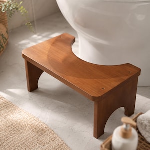 May include: A brown wooden toilet stool featuring a U-shaped cutout designed to fit around the base of a toilet. The stool has a smooth, flat top surface with four visible screw heads and arched cutouts on the two side support legs.