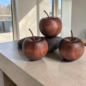 May include: Three decorative apple sculptures carved from dark brown wood, each featuring a smooth, rounded body and a thin, upright stem. The apples are arranged with two positioned in the front and one elevated on a dark, smooth stone base.