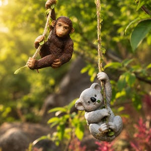 May include: A brown chimpanzee figurine and a grey koala figurine, each hanging from a separate vertical rope adorned with small green leaves. The chimpanzee is positioned above the koala, and both figures are sculpted with textured fur.