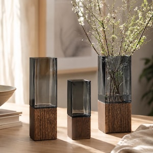 May include: Three rectangular vases featuring dark gray glass tops and solid wood bases. The vases vary in height, with the tallest one on the right containing thin branches with small white flowers. The glass is translucent and smooth.