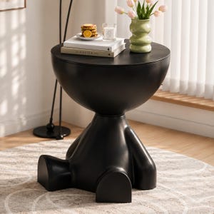 Puede incluir: Una mesa auxiliar negra mate con una superficie redondeada y una base con forma de figura sentada. Sobre la mesa hay dos libros apilados, un plato pequeño con gofres y rodajas de naranja, una vela en vaso y un jarrón de color verde claro.