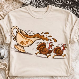 May include: A cream-colored short-sleeve t-shirt featuring a graphic illustration of a gravy boat pouring brown gravy onto two running brown turkeys with red heads and orange beaks, depicted in a cartoon style with motion lines.