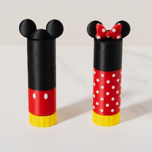 May include: Two cylindrical containers with character-themed caps featuring black mouse ears. One container is black and red with two white ovals, while the other is black and red with white polka dots and a red bow with white dots. Both have yellow bases.